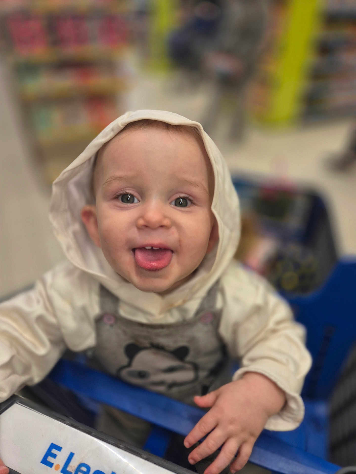 Elyo a rejoint le concours — aidez-le/la à gagner de superbes lots ! baby, child, shopping_cart, hood, clothing, face, tongue_out, smile, hand, store, blurred_background, infant, cute, person, indoor, panda, hooded_jacket, cart, playful, eyes