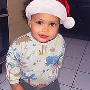 Adriel participe au concours pour gagner de l'argent avec cette photo : brown_socks, child, cute, dinosaur_print, festive, footwear, holiday, holiday_season, indoor, kitchen, looking_up, pants, person, santa_hat, smiling, standing, sweater, tile_floor, toddler, young_child
