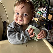 Ezio participe au concours pour gagner de l'argent avec cette photo : baby, toddler, highchair, messy_face, crumbs, cup, hands, blue_eyes, gray_sweater, hair, tray, indoor, plant, smile, portrait, closeup, food_scraps, chair, feeding_time, cute
