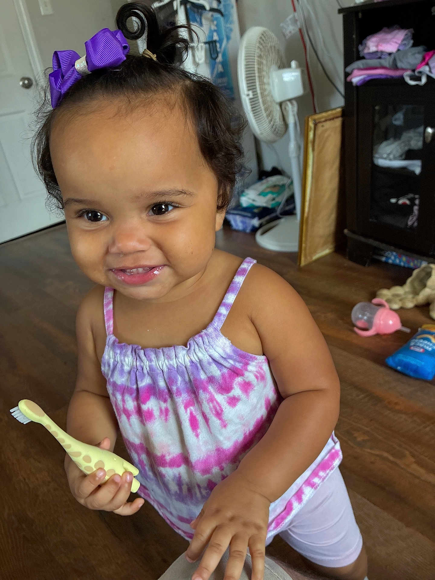 Kyomi is registered to the contest to win money with this photo: toddler, child, smiling, purple_bow, tie_dye, toothbrush, giraffe_shaped, indoor, wooden_floor, cabinet, fan, clothing, person, happy, cute, hair, hand, face, baby, playful