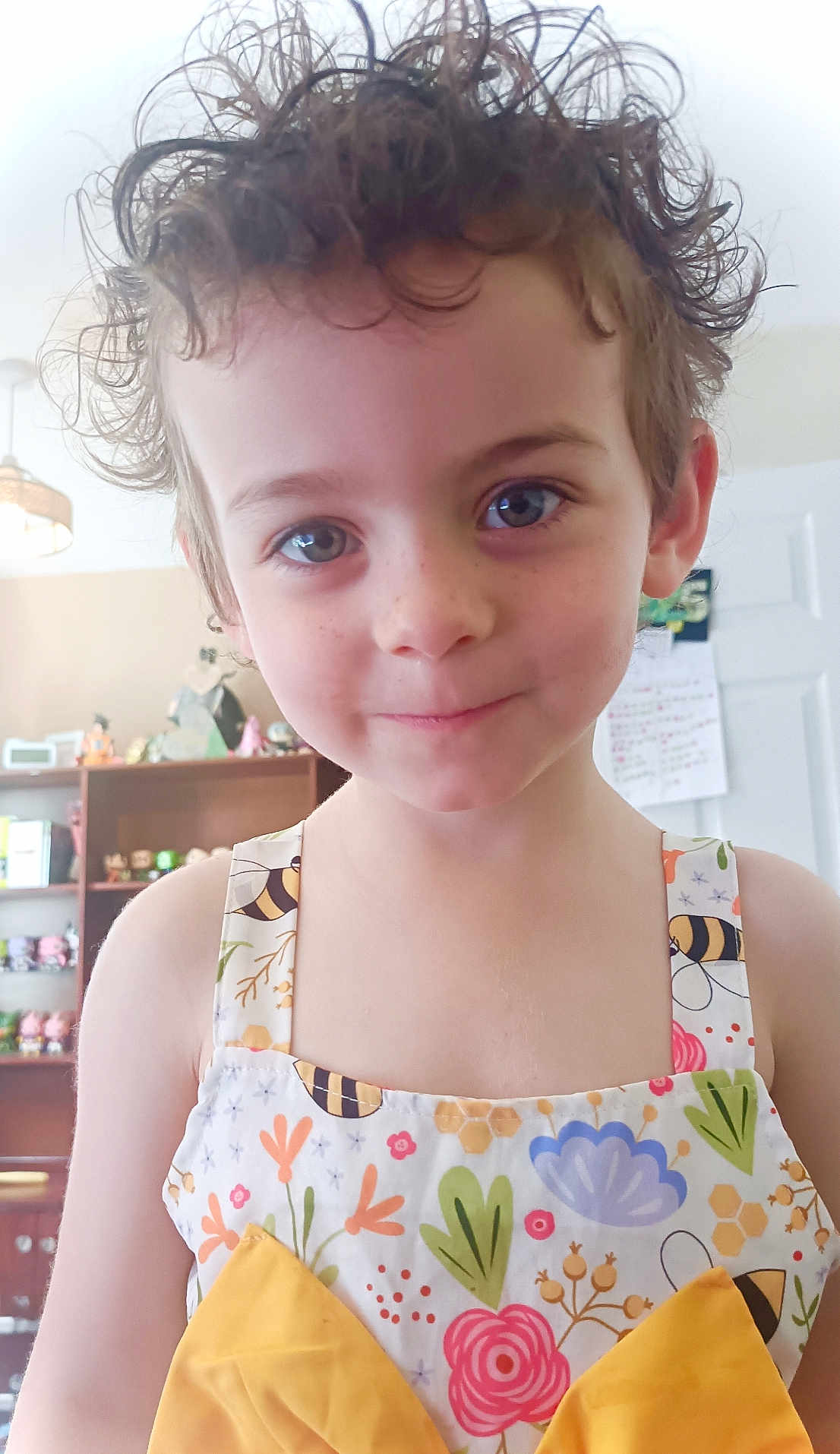 Ruby is registered to the contest to win money with this photo: child, curly_hair, blue_eyes, smiling, floral_dress, yellow_bow, indoor, toys, shelves, background, person, cute, portrait, freckles, young, colorful, happy, fashion, hair, face