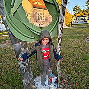 Adrian is registered to the contest to win money with this photo: child, outdoor, autumn, sculpture, leaf_sculpture, playground, standing, hooded_jacket, pants, sneakers, grass, trees, houses, neighborhood, portrait, full_body, architecture, blue_sky, daylight, playful