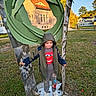 child, outdoor, autumn, sculpture, leaf_sculpture, playground, standing, hooded_jacket, pants, sneakers, grass, trees, houses, neighborhood, portrait, full_body, architecture, blue_sky, daylight, playful
