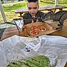 child, toy_dinosaur, pepperoni, cracker, picnic_table, outdoor, grass, house, drink_pouch, wood, paper_wrapper, food, snack, curly_hair, daylight, bench, playful, greenery, casual, portrait