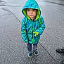 boy, casual_clothing, child, daylight, dinosaur_pattern, expressionless, footwear, grass, hood, outdoor, pavement, person, rain_jacket, road, shoes, standing, street, toy, weather, wet_surface
