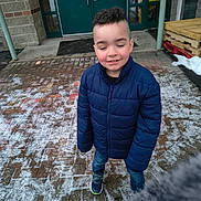 Adrian is registered to the contest to win money with this photo: child, boy, jacket, blue_jacket, closed_eyes, smiling, outdoor, snow, brick_pavement, door, green_door, wooden_pallets, casual, winter_clothing, sneakers, sidewalk, concrete, building_entrance, person, young