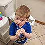 Benjamin is registered to the contest to win money with this photo: child, toothbrush, bathroom, toilet, tile_floor, blue_shirt, shoes, person, young_child, brushing_teeth, storage_drawer, floor, head, hair, necklace, wall, home_interior, daily_life, indoors, expression
