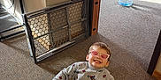 Benjamin is registered to the contest to win money with this photo: child, indoor, tricycle, smiling, sunglasses, grey_outfit, motorcycle_pattern, american_flag, carpet, baby_gate, toy, stuffed_animal, playroom, happy, person, floor, window_light, casual_clothing, small_bike, pink_accessory