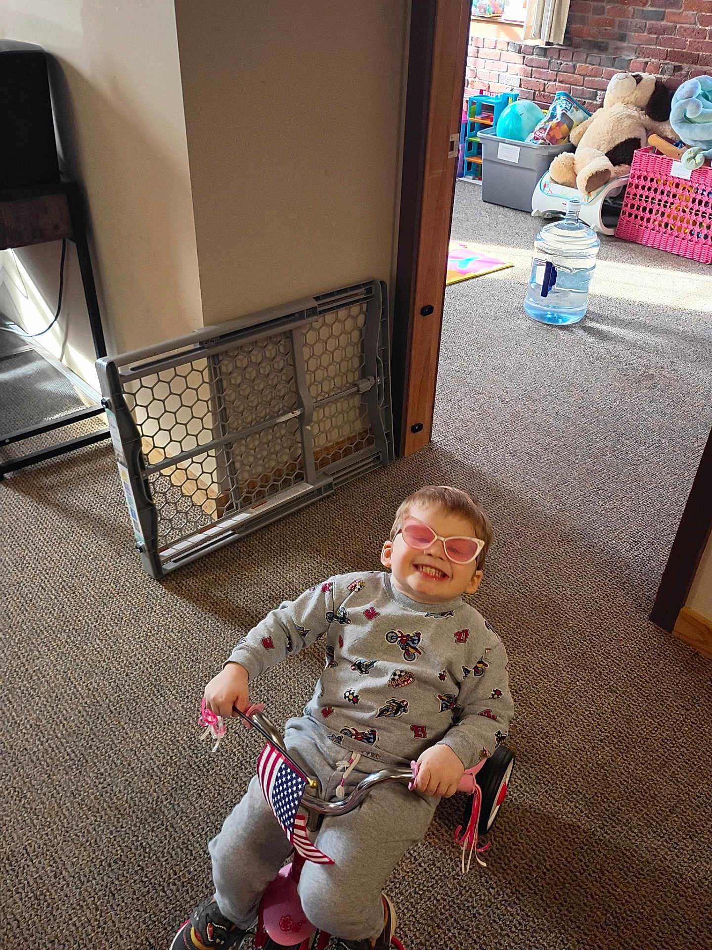 Benjamin is registered to the contest to win money with this photo: child, indoor, tricycle, smiling, sunglasses, grey_outfit, motorcycle_pattern, american_flag, carpet, baby_gate, toy, stuffed_animal, playroom, happy, person, floor, window_light, casual_clothing, small_bike, pink_accessory