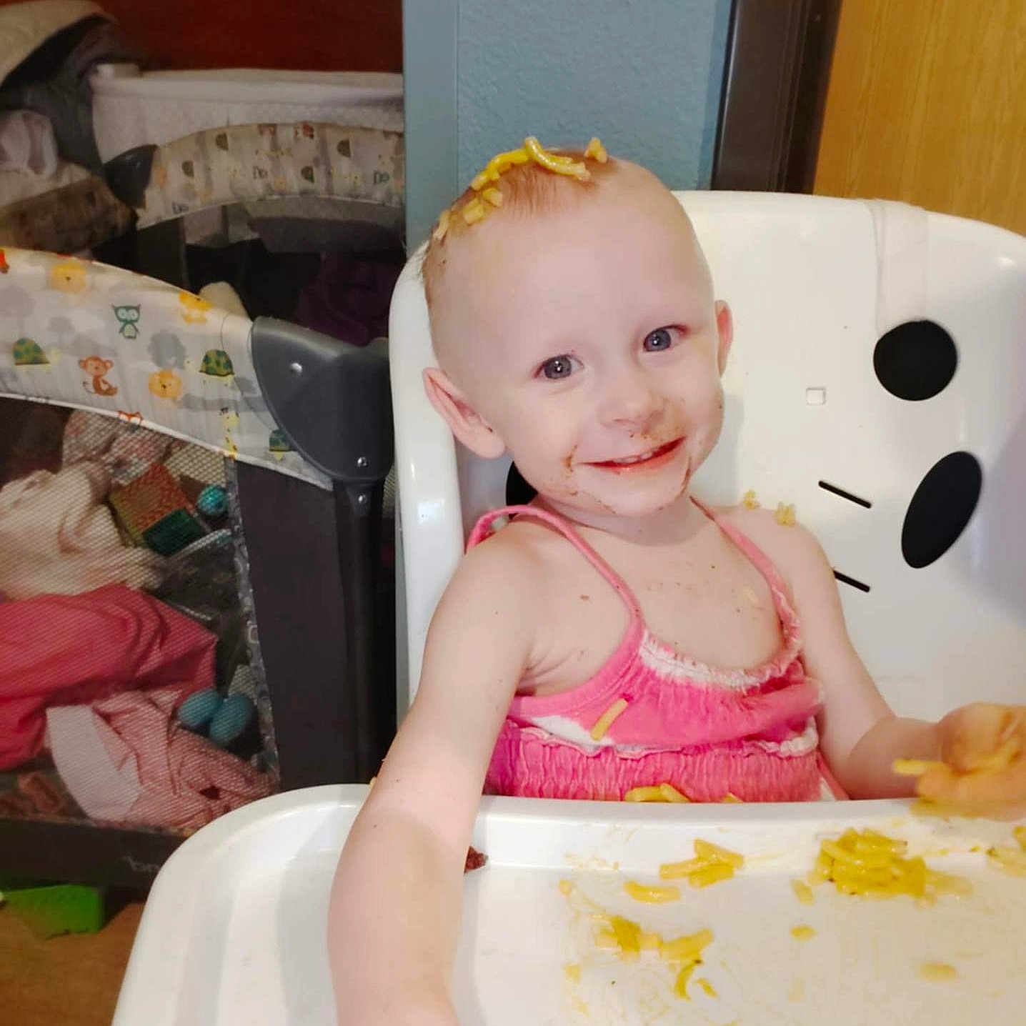 Schela is registered to the contest to win money with this photo: blue_eyes, chair, child, cute, face, food, fun, hair, hand, high_chair, indoor, macaroni, meal_time, messy, pink_dress, playpen, skin, smiling, table, toddler