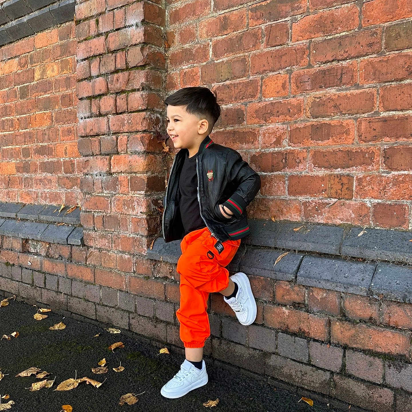 Matthias-Gabriel is registered to the contest to win money with this photo: autumn_leaves, black_jacket, boy, brick_wall, casual_pose, child, cool, daylight, fall, fashion, happy, orange_pants, outdoor, side_view, smiling, standing, street_style, urban, white_sneakers, young_person