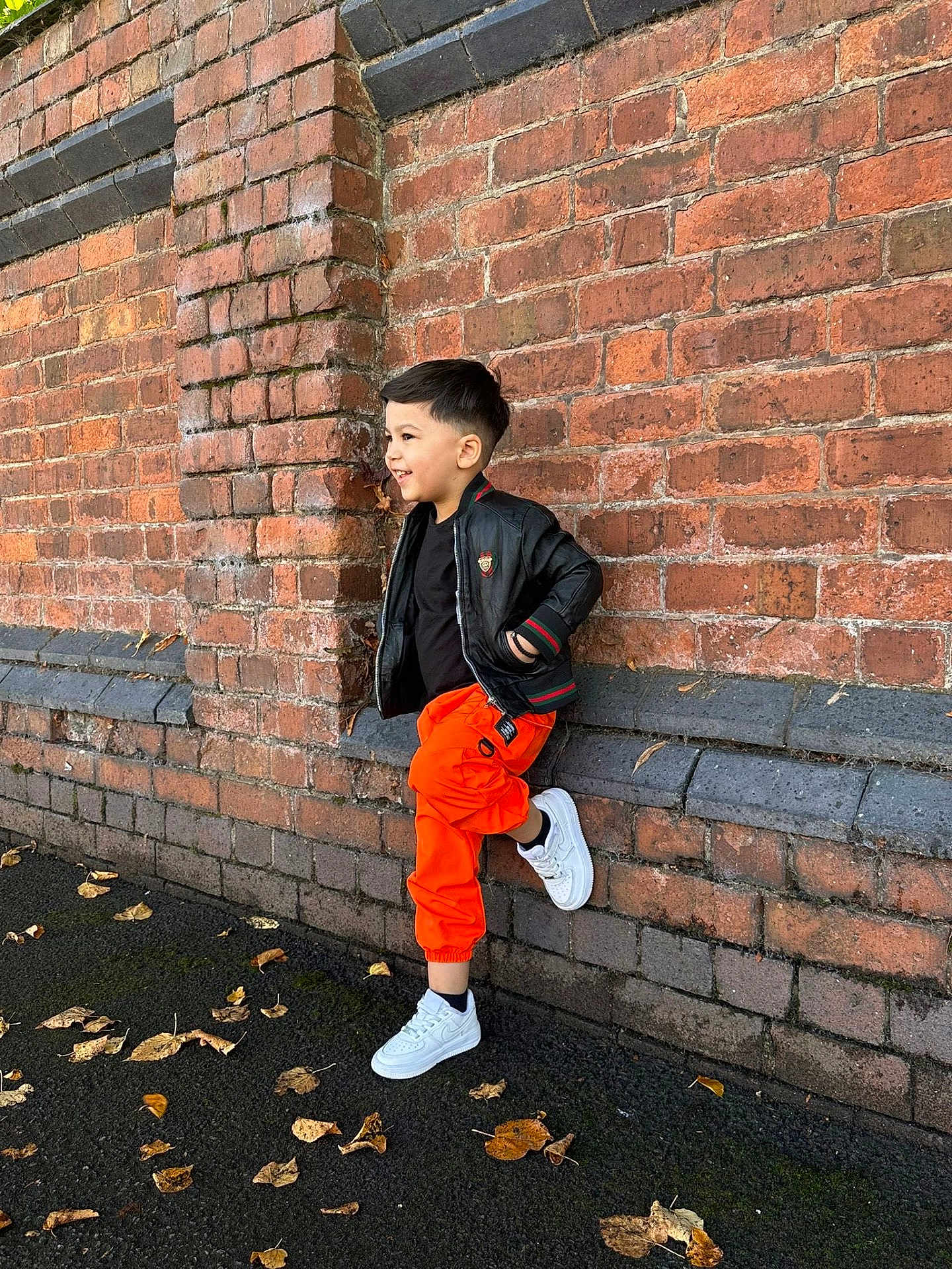 Matthias-Gabriel is registered to the contest to win money with this photo: child, boy, brick_wall, orange_pants, black_jacket, white_sneakers, side_view, smiling, autumn_leaves, outdoor, casual_pose, street_style, young_person, urban, fashion, standing, fall, daylight, happy, cool
