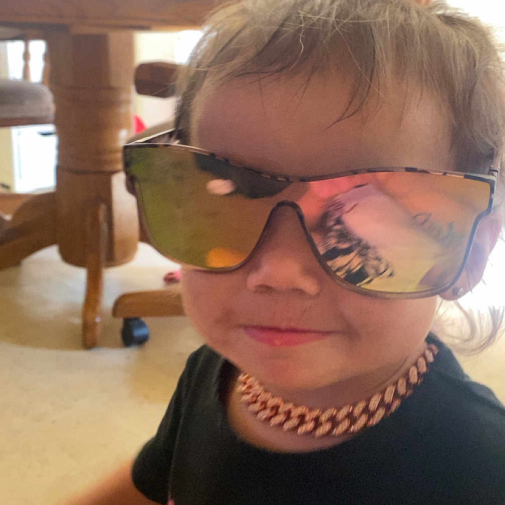 Alyna is registered to the contest to win money with this photo: accessory, black_shirt, casual_clothing, child, cute, face, floor, hair, indoor, kitchen, necklace, person, portrait, reflection, smile, sunglasses, table, toddler, wooden_chair, young_child