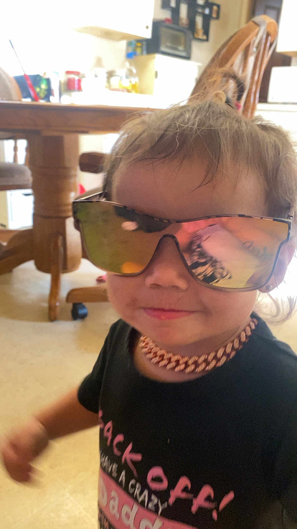 Alyna is registered to the contest to win money with this photo: toddler, child, sunglasses, necklace, black_shirt, indoor, kitchen, wooden_chair, table, reflection, smile, person, young_child, casual_clothing, floor, hair, accessory, face, cute, portrait