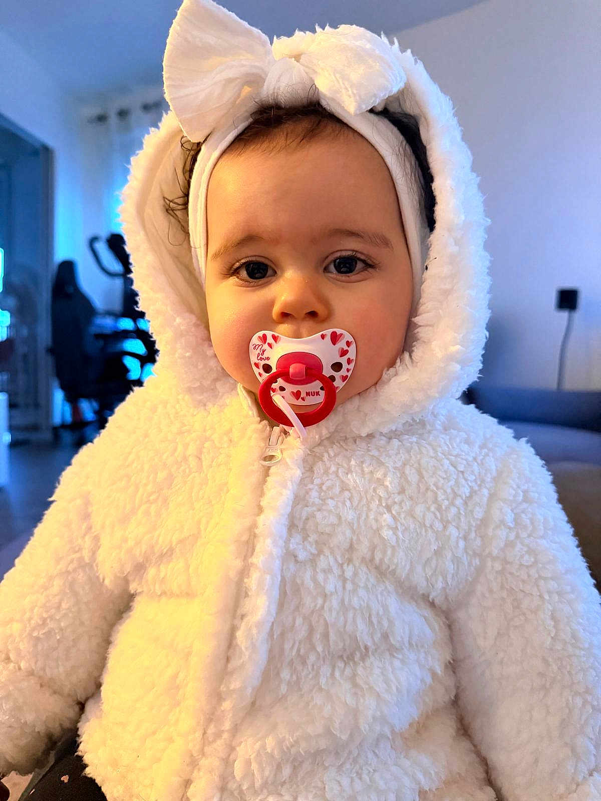 Julia a rejoint le concours — aidez-le/la à gagner de superbes lots ! baby, pacifier, white_jacket, headband, bow, indoor, child, cute, fluffy_clothing, infant, face, person, young_child, warm_clothing, portrait, sitting, cozy, room, soft_light, expressionless