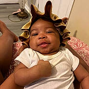 Josiah is registered to the contest to win money with this photo: baby, infant, smile, bonnet, white_clothing, arm, hand, person, indoor, lamp, floor, blanket, pattern, relaxing, cute, skin, face, headwear, comfort, home