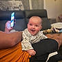baby, child, smiling, happy, mickey_mouse, shirt, person, phone, hand, couch, living_room, blurred_background, arm, orange_shirt, black_shorts, indoor, infant, cute, joyful, portrait
