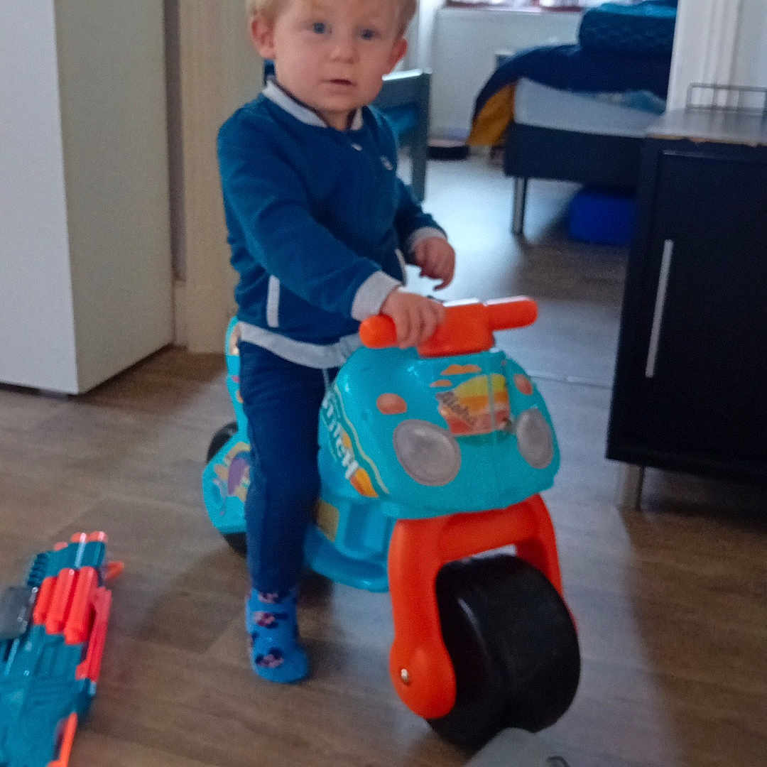 Noa a rejoint le concours — aidez-le/la à gagner de superbes lots ! blue_clothing, cabinet, child, curious, floor_toys, furniture, home, indoor, natural_light, person, plastic_toy, playful, ride_on_toy, room, socks, standing, toddler, toy, window, wooden_floor