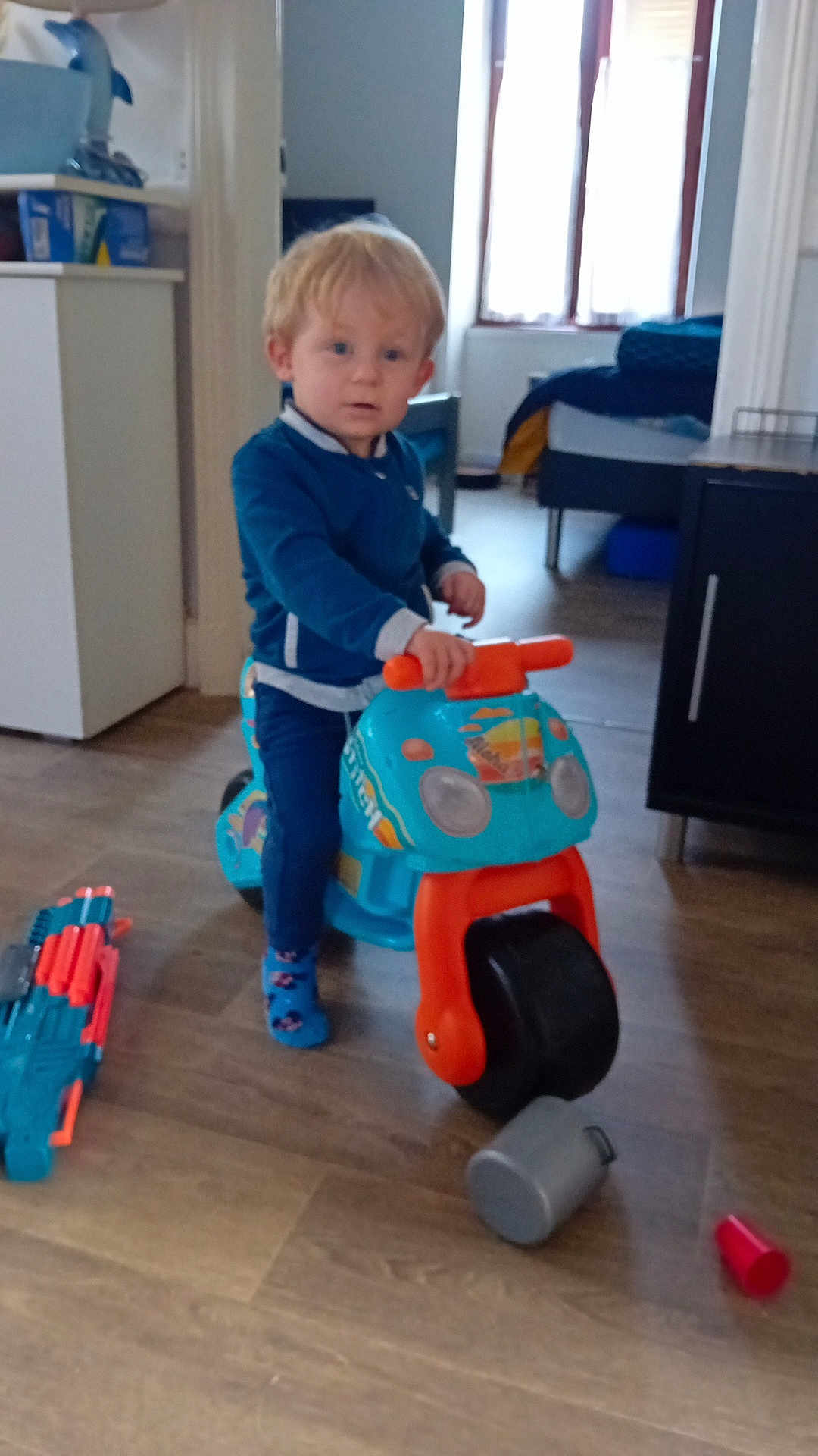 Noa a rejoint le concours — aidez-le/la à gagner de superbes lots ! toddler, child, indoor, toy, plastic_toy, ride_on_toy, blue_clothing, socks, wooden_floor, window, furniture, cabinet, floor_toys, curious, standing, person, home, natural_light, room, playful