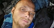 Apollo is registered to the contest to win money with this photo: beard, black_dog, blue_shirt, casual, close_up, clothing, dog, face, furniture, head, human, indoor, man, pet, pile_of_clothes, portrait, relaxed, resting, snuggling, white_markings