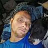 man, dog, face, portrait, indoor, casual, clothing, blue_shirt, close_up, snuggling, black_dog, white_markings, relaxed, beard, head, furniture, pile_of_clothes, resting, pet, human