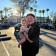 Daniella is registered to the contest to win money with this photo: adult, baby, child, tattoo, glasses, smile, outdoor, parking_lot, car, palm_tree, sunlight, tree, person, holding, happy, casual_clothing, daytime, family, urban, portrait