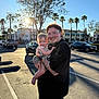 adult, baby, child, tattoo, glasses, smile, outdoor, parking_lot, car, palm_tree, sunlight, tree, person, holding, happy, casual_clothing, daytime, family, urban, portrait