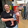 Daniella is registered to the contest to win money with this photo: woman, baby, child, holding, mini_golf_club, outdoor, amusement_park, sign, building, clothing, shorts, tshirt, footwear, smile, happy, person, playful, toy, recreation, daytime