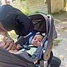 baby, child, stroller, car_seat, person, adult, outdoor, greenery, hair_clips, infant, seatbelt, sidewalk, building, sunlight, shade, clothing, hand, portrait, closeup, park