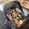 baby, car_seat, child, infant, yellow_clothing, hair_clips, indoor, floor, metal_rack, box, straps, footwear, curious_expression, person, seat, casual, black_hair, light, toddler, safety