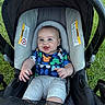 Asher is registered to the contest to win money with this photo: baby, infant, child, car_seat, stroller, smiling, happy, outdoor, grass, portrait, sitting, lying_down, colorful_clothing, blue_shirt, gray_pants, baby_carrier, canopy, safety_labels, hands, face