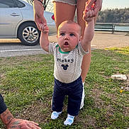 Asher is registered to the contest to win money with this photo: baby, child, infant, outdoor, park, grass, lakeside, adult, parent, holding_hands, standing, smiling, surprised, portrait, mickey_mouse, clothing, socks, car, trees, sunlight