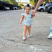 Lara is registered to the contest to win money with this photo: toddler, child, person, walking, hand_holding, romper, sandals, sidewalk, street, cars, trees, daylight, blue_sky, outdoor, concrete, crack, adult, clothing, footwear, support