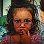 child, curly_hair, glasses, heart_shaped_glasses, finger_in_mouth, thoughtful, portrait, indoor, pink_striped_shirt, close_up, face, person, young, casual, expression, cute, eyewear, fashion, childhood, skin