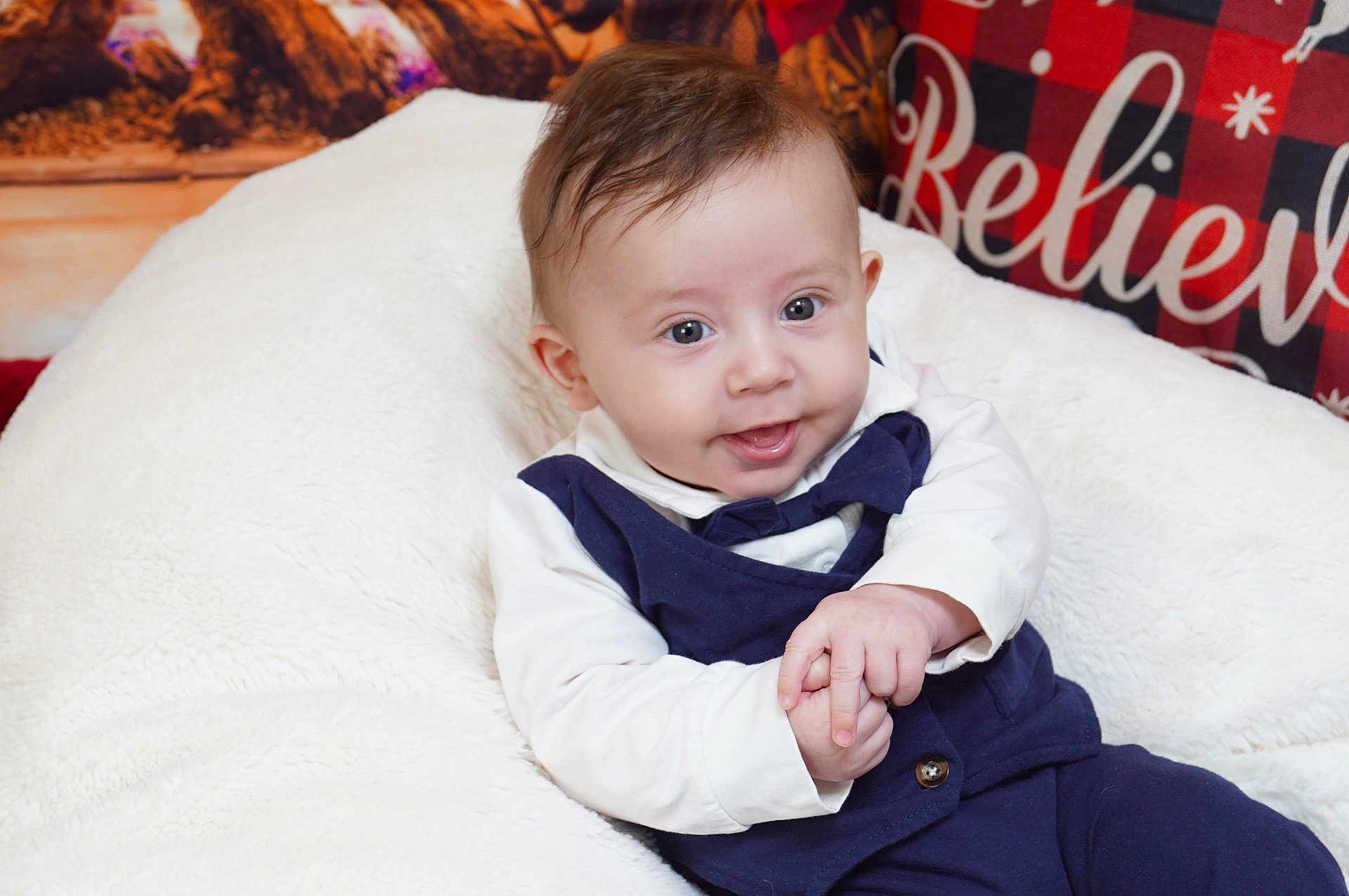 Jordan is registered to the contest to win money with this photo: baby, infant, smiling, hands, white_cushion, navy_outfit, bow_tie, face, eyes, hair, indoor, pillow, plaid_pattern, text, believe, cozy, festive, child, portrait, cute