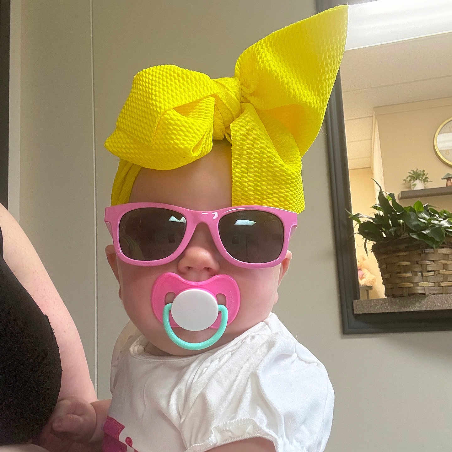 Jennalynn is registered to the contest to win money with this photo: baby, child, sunglasses, pacifier, yellow_bow, white_shirt, indoor, person, fashion, accessory, cute, infant, headwear, pink, holding, plant, mirror, reflection, adult_arm, casual