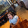 child, toddler, laughing, high_chair, wooden_table, hair_clips, indoor, restaurant, mural, mountains, smiling, short_hair, blue_shirt, daisies, earrings, chair, floor, table, furniture, happy