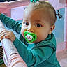 toddler, child, pacifier, playpen, pink, teal_shirt, pigtails, indoor, toy_box, cartoon, curious, baby, face, hand, standing, furniture, colorful, young_child, expression, hair