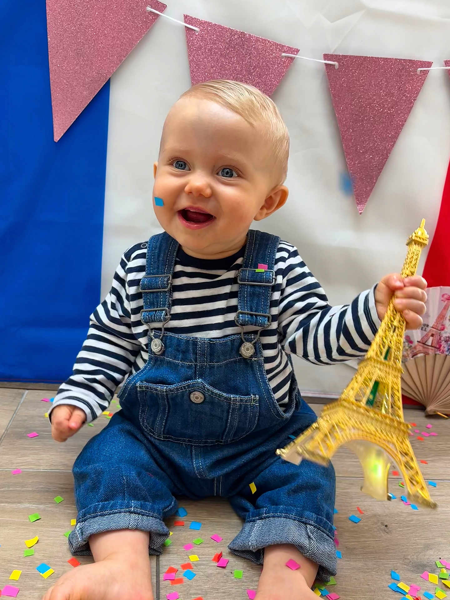 Ewen a rejoint le concours — aidez-le/la à gagner de superbes lots ! toddler, child, denim_overalls, striped_shirt, confetti, floor, toy, eiffel_tower, smiling, blue_eyes, pink_banners, glitter, celebration, indoor, cute, happy, portrait, sitting, barefoot, festive