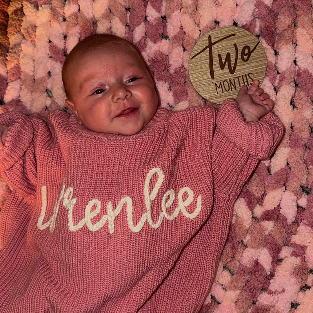 Wrenlee Jade is registered to the contest to win money with this photo: baby, infant, two_months, blanket, knit, pink, sweater, person, face, smile, hand, wooden_sign, milestone, cute, lying_down, soft_texture, cozy, indoors, child, newborn