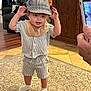Jayce is registered to the contest to win money with this photo: toddler, child, striped_clothing, hat, socks, carpet, living_room, furniture, armchair, pillow, cat_design, wooden_cabinet, smartphone, hand, floor_tiles, baby_bib, smiling, standing, indoor, cute
