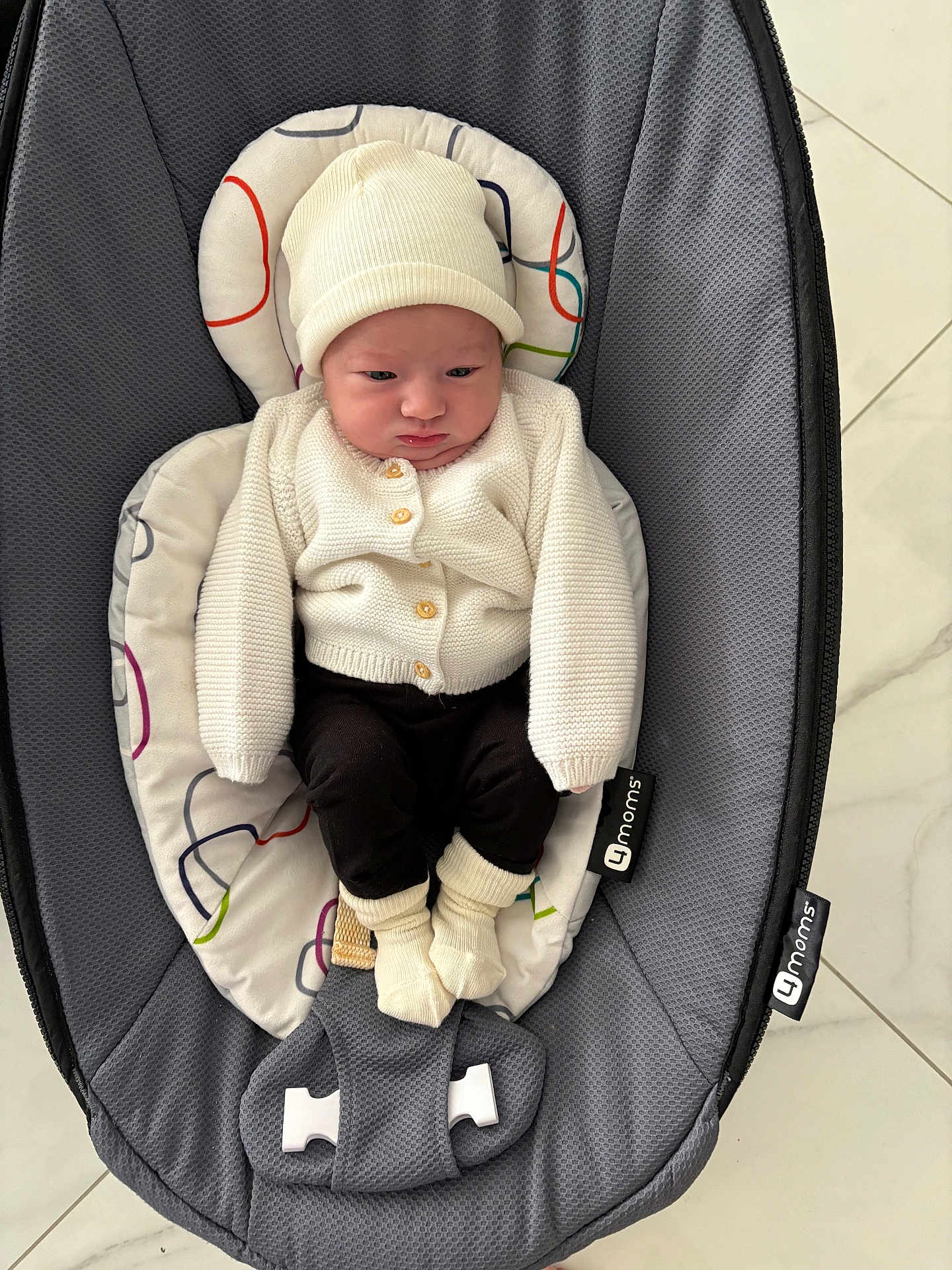 Mila a rejoint le concours — aidez-le/la à gagner de superbes lots ! baby, infant, hat, sweater, baby_rocker, gray, white, black_pants, socks, indoor, floor, tile_floor, seated, child, cute, cozy, clothing, newborn, resting, serious_expression