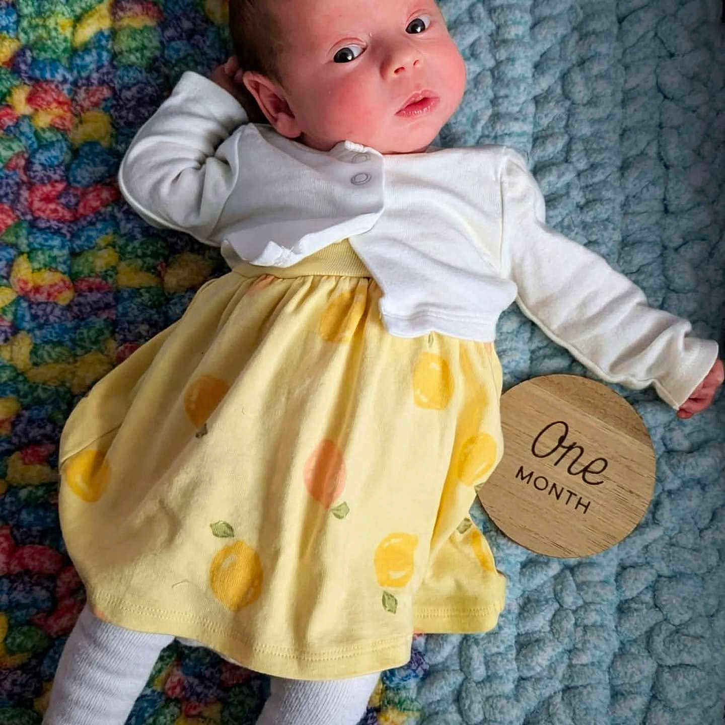 Malia is registered to the contest to win money with this photo: baby, infant, one_month, blanket, knitted_blanket, yellow_dress, white_cardigan, face, person, lying_down, milestone_sign, wooden_sign, cute, newborn, child, colorful, patterned, indoors, soft_texture, clothing