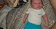 Elias joined the competition — help win amazing prizes! baby, infant, child, portrait, lying_down, bed, bedroom, blanket, pillow, patterned_bedding, white_shirt, turquoise_shorts, nike, sitting, face, eyes_open, calm, indoor, adult_arm, cozy