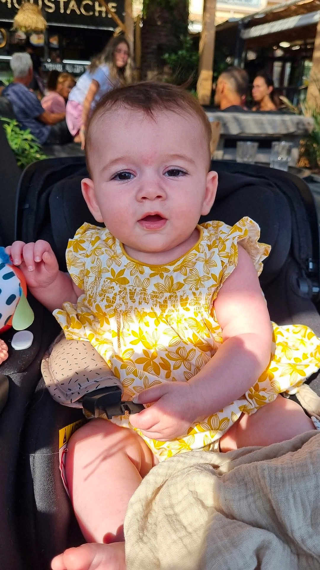 Ellvyra participe au concours pour gagner de l'argent avec cette photo : baby, blurred_background, cafe, car_seat, casual, child, curious_expression, cute, daylight, infant, outdoor, people, person, portrait, seatbelt, smile, summer_clothing, sunlight, toy, yellow_dress