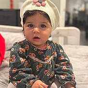 Amira is registered to the contest to win money with this photo: baby, child, toddler, portrait, sitting, indoor, floral_dress, knit_hat, flower_hat, wide_eyes, open_mouth, dark_hair, smiling, clothing, couch, home, soft_lighting, blurred_background, snack, cute