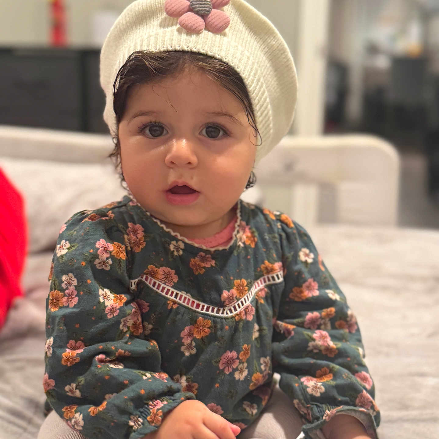 Amira is registered to the contest to win money with this photo: baby, child, toddler, portrait, sitting, indoor, floral_dress, knit_hat, flower_hat, wide_eyes, open_mouth, dark_hair, smiling, clothing, couch, home, soft_lighting, blurred_background, snack, cute