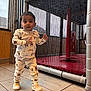 child, toddler, indoor, play_area, mesh_net, red_mat, tile_floor, pajamas, sneakers, standing, curious_expression, face, full_body, window, wood_panel, safety_padding, vertical_poles, netting, candid, playground_equipment
