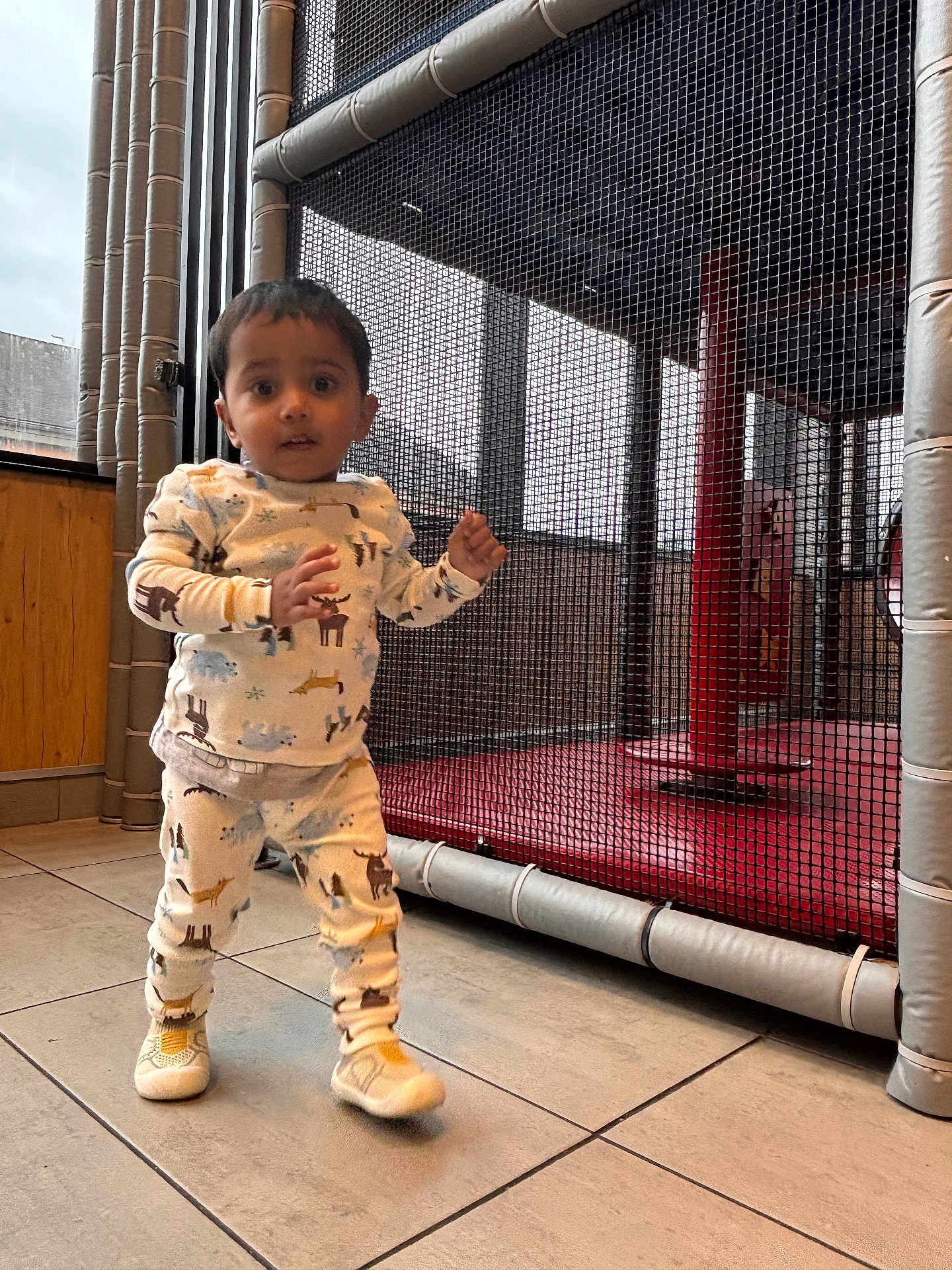 Tanush is registered to the contest to win money with this photo: child, toddler, indoor, play_area, mesh_net, red_mat, tile_floor, pajamas, sneakers, standing, curious_expression, face, full_body, window, wood_panel, safety_padding, vertical_poles, netting, candid, playground_equipment