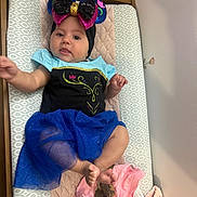 Gianna joined the competition — help win amazing prizes! baby, infant, headband, blue_skirt, black_top, changing_pad, blanket, clothing, cute, child, person, indoor, soft_texture, pink_clothing, sequin_bow, reaching_hand, tongue_out, foot, lying_down, wooden_frame
